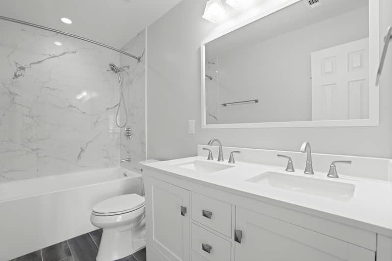 Hall bathroom with a white dual-sink vanity and quartz counter, large mirror, toilet, and a tub-shower clad in marble-look tile