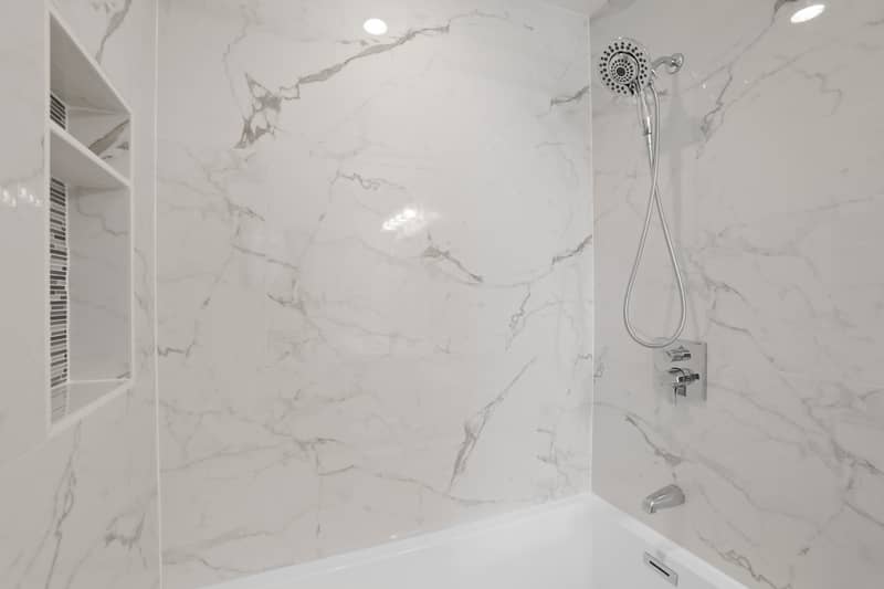 Close-up of a tub-shower surround in glossy marble-look tile with a recessed shelving niche, handheld showerhead, and chrome fixtures
