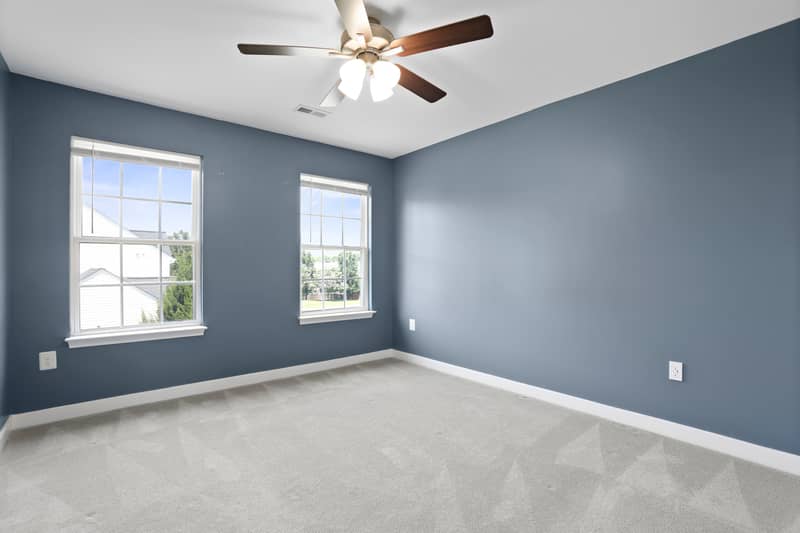 Slate-blue carpeted bedroom corner with a lighted ceiling fan and two windows looking onto the yard