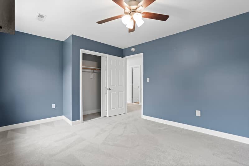 Bedroom with soft blue-gray walls, plush light gray carpet, a wood-blade ceiling fan, an open reach-in closet with hanging rod, and a doorway leading to the carpeted hallway