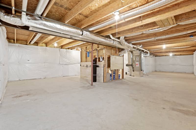Wide view of an unfinished basement with smooth concrete floor, exposed wood joists and insulated ductwork overhead, white vapor-barrier walls, and mechanical equipment in the center