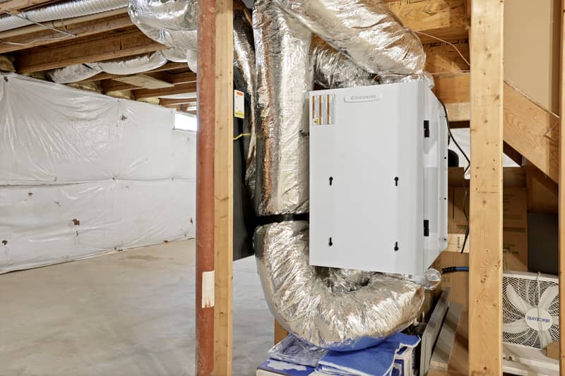 Close view of a white wall-mounted HVAC air handler unit connected to insulated flexible ducting amid exposed wood framing in the basement