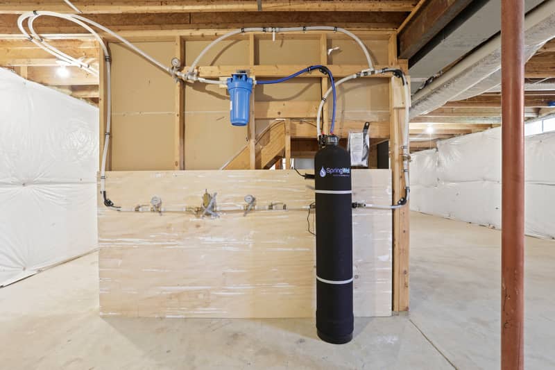 Basement utility area showing a SpringWell whole-house water filtration and softening system with plumbing manifold mounted on a plywood backing