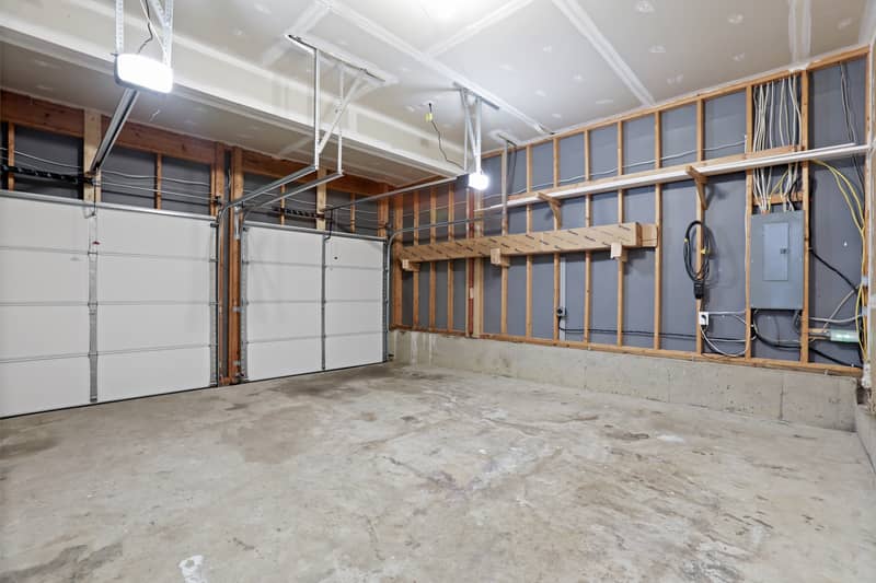 Interior of a two-car garage with two white sectional doors, automatic openers, concrete floor, exposed wall framing, and an electrical panel with overhead lumber storage rack